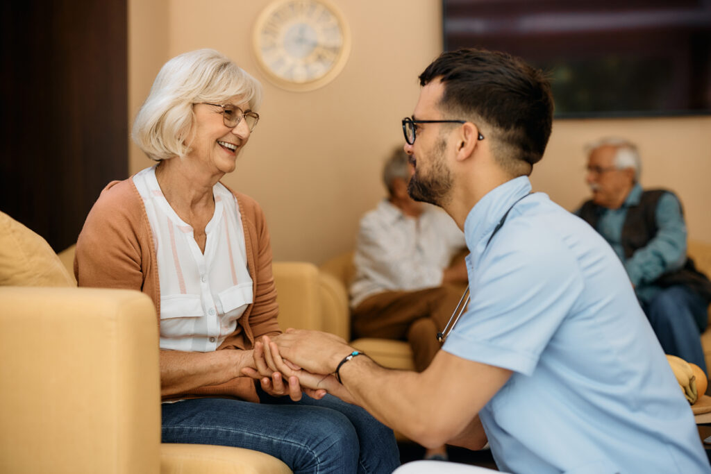 Happy male caregiver and senior woman hold hands while communicating at residential care home.